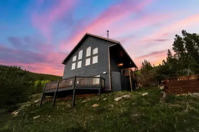 a black house with a porch on a hill with a sunset at Family Mountain Getaway w Hot Tub and Amazing Views! Close to Hiking & Skiing in Idaho Springs